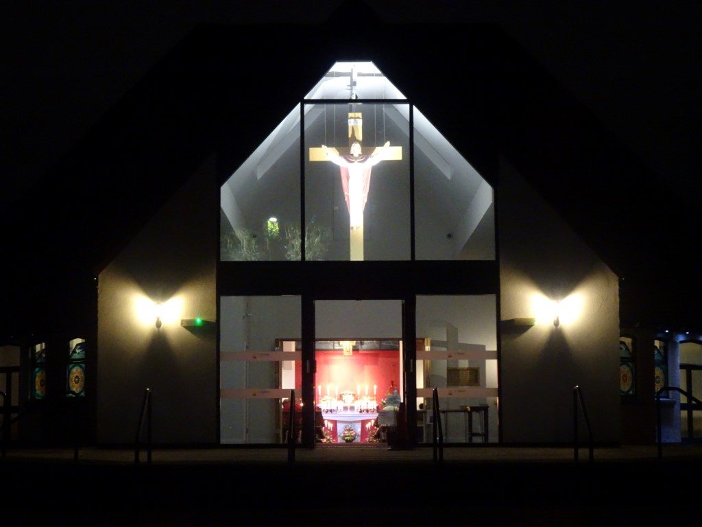 St Clare's Church lit at night, with the sanctuary crucifix visible through the glass gable